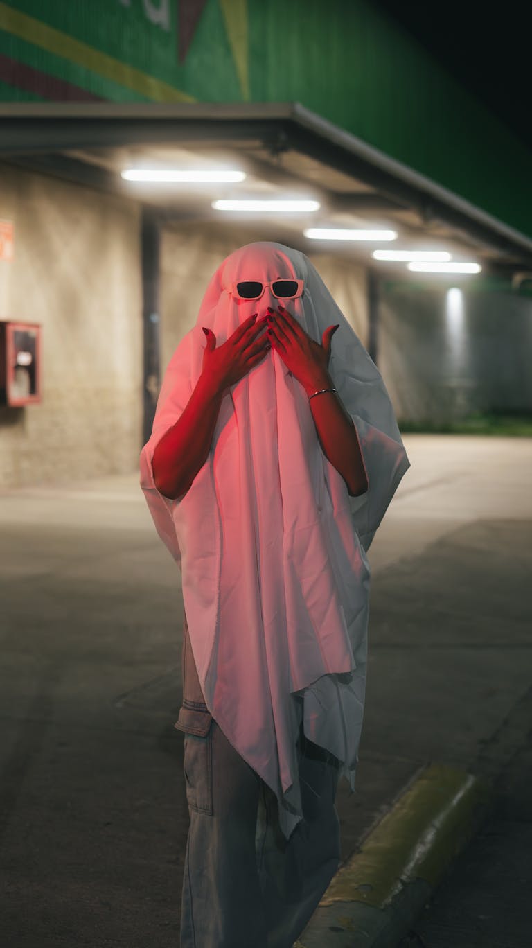 A person in a ghost costume with sunglasses poses outdoors at night, creating a spooky yet humorous scene.